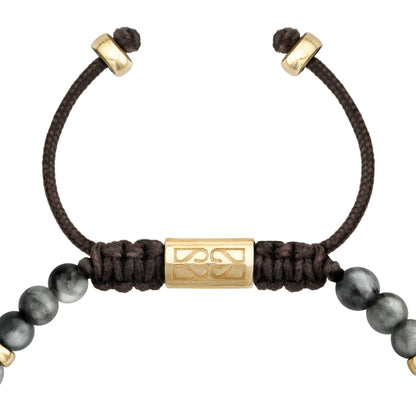 Detalle del cierre ajustable de cordón negro con un cilindro dorado grabado, en una pulsera de cuentas grises y detalles dorados.