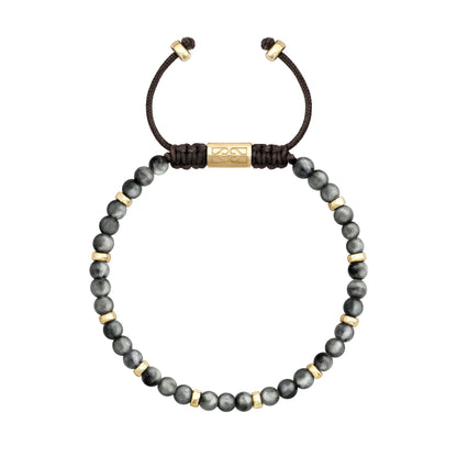 Pulsera de cuentas grises con separadores dorados, cierre ajustable de cordón negro y un cilindro dorado grabado en el centro.