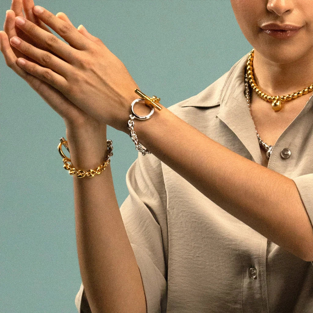 Persona luciendo pulsera de eslabones dorados y plateados con cierre de barra en forma de Omega, acompañada de un collar dorado. - Joyería y bisutería fina